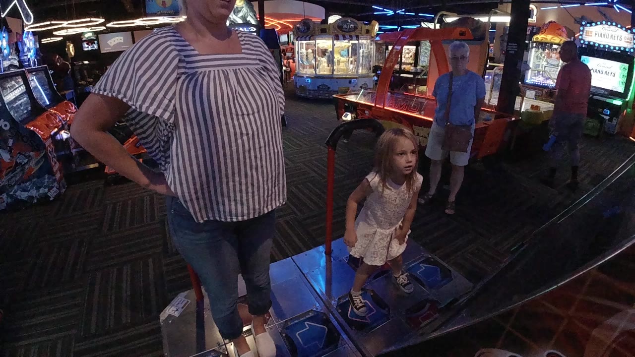 In Denver, a woman stands at the left edge while a little girl faces the camera near a blue-lit arcade dance platform with game machines behind them.