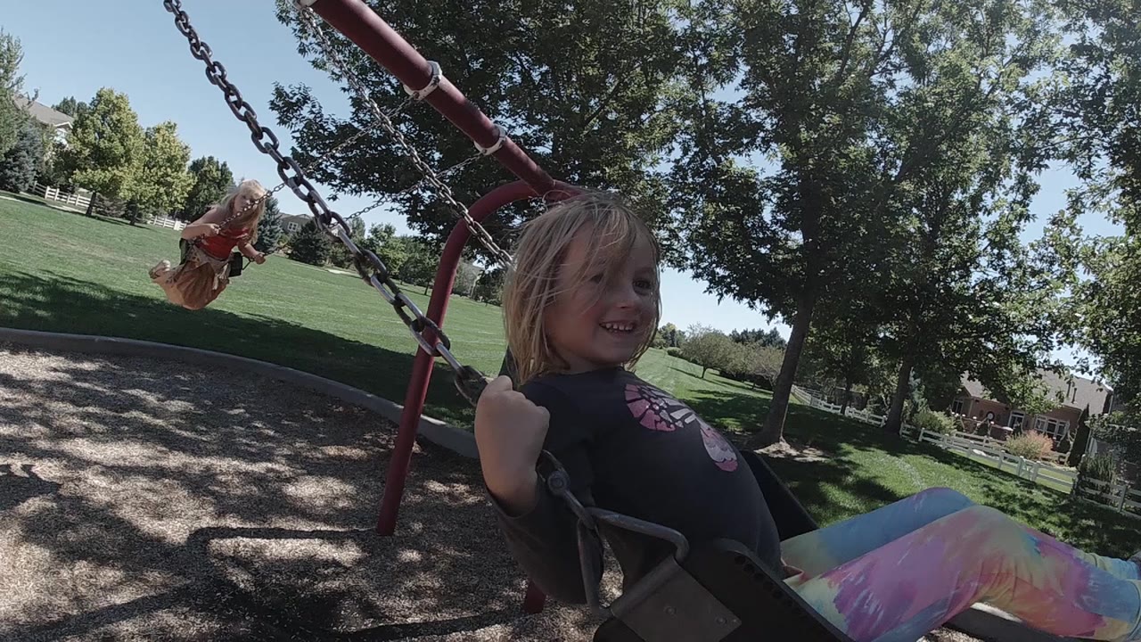 The same girl remains close in the swing seat, smiling beside dark swing chains while a second child rides another swing in the sunny Denver park background.
