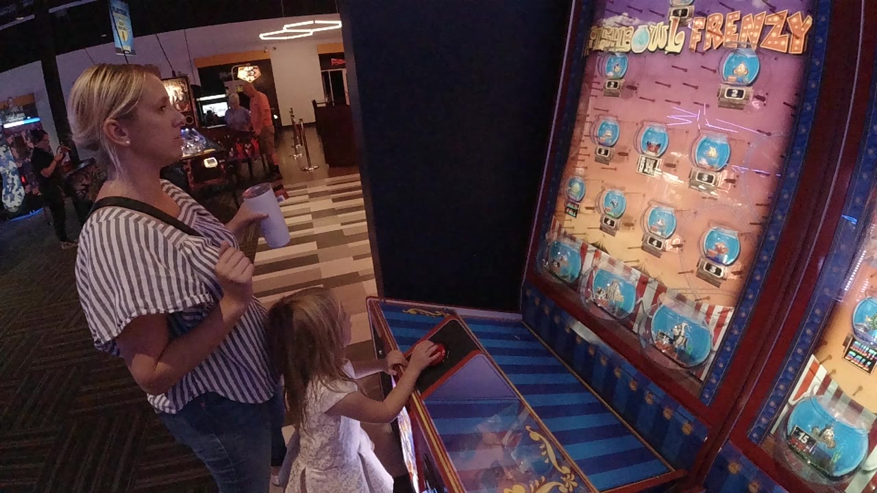 At 0:00 in Denver, a blonde woman and young girl stand beside a tall illustrated arcade game with blue play panels and red trim.