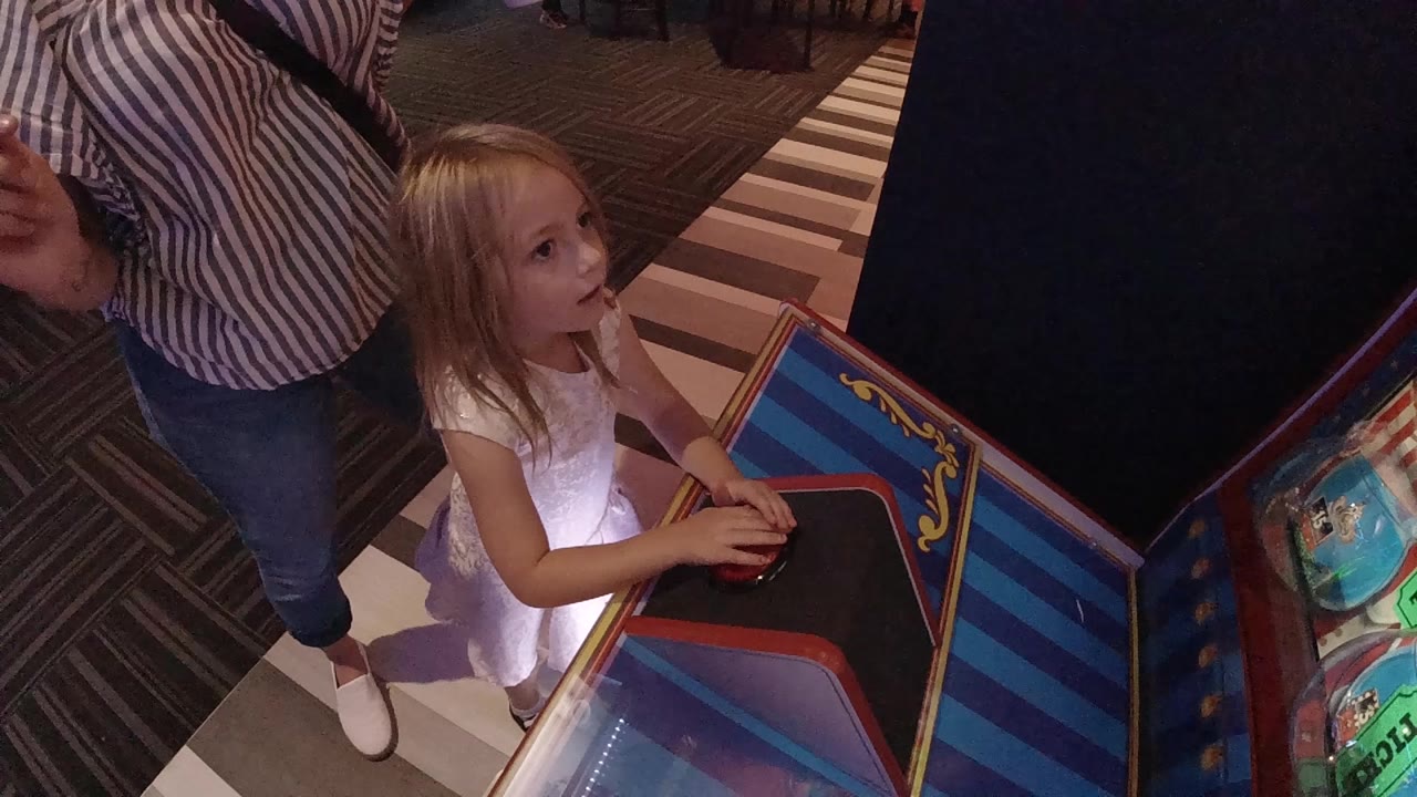 At 0:01 in Denver, the girl looks toward the arcade screen area with both hands on the dark control panel and the adult's striped shirt visible to the left.