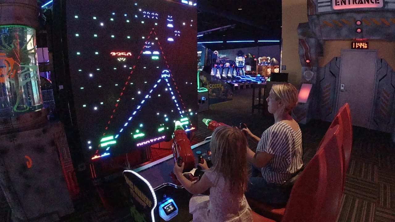 In Colorado, a mother and daughter sit at a glowing arcade shooter cabinet as the game screen shows rows of colored lights near the start of play.