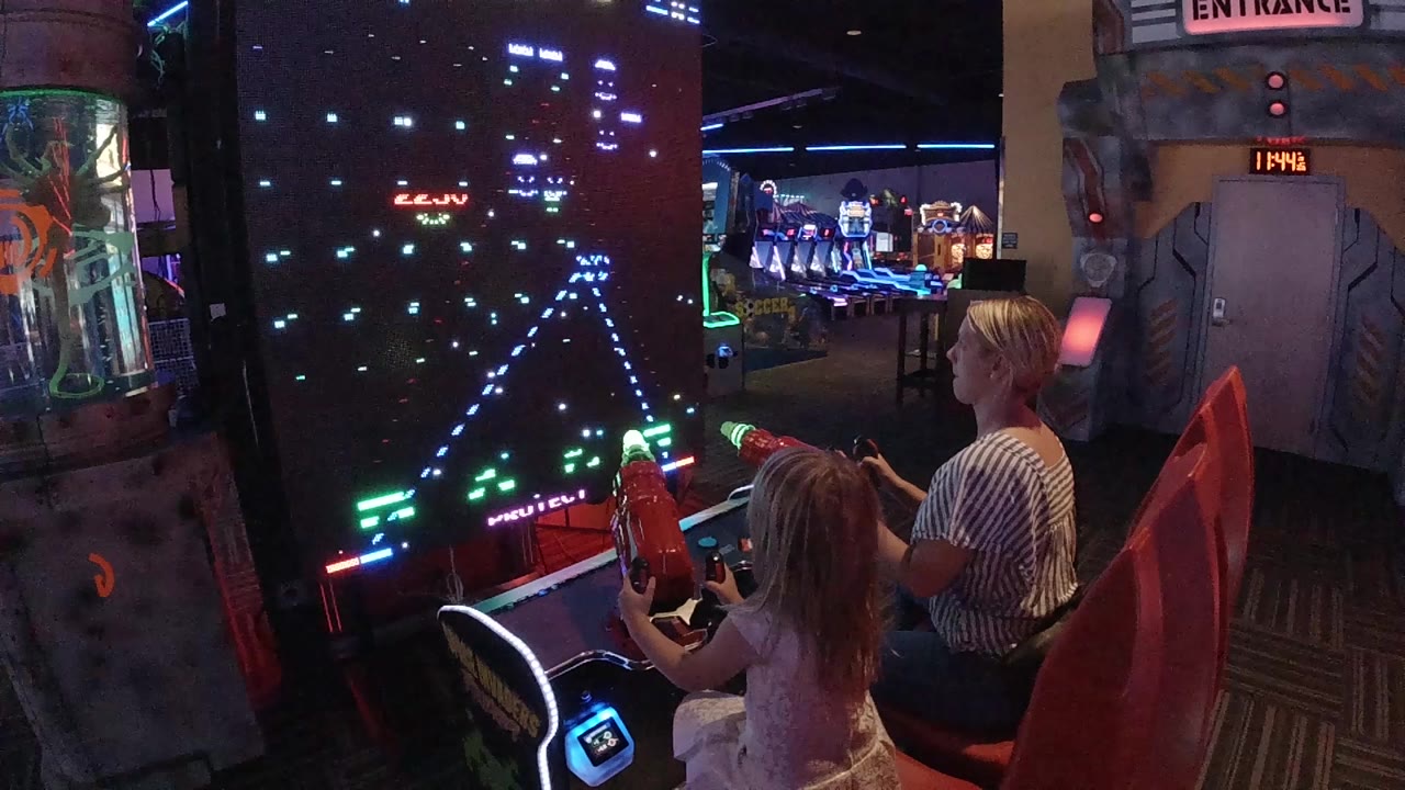 The girl grips the front red control while the woman sits beside her, both facing a tall black game screen filled with blue and green pixel targets.