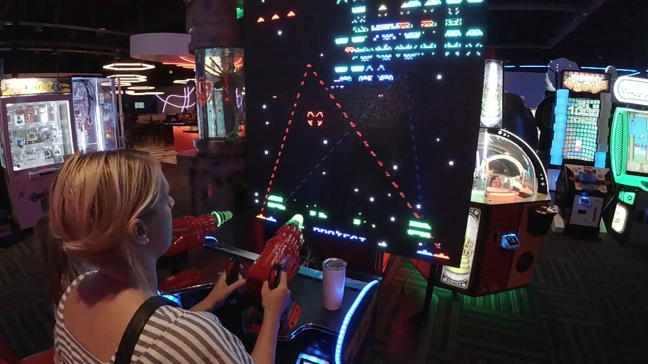 A blonde woman in Colorado plays a Galaga arcade cabinet from a close rear angle, with red controls in her hands and other glowing machines behind the screen.