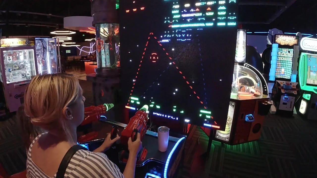 The woman continues playing Galaga in Colorado, with the game screen showing bright triangular attack paths and the arcade machines glowing around the cabinet.