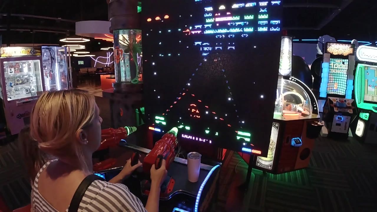 A side rear view shows the woman's head and shoulders near the red control panel while Galaga enemies and colored lights fill the tall screen.