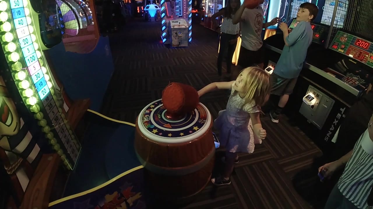 The red mallet drops onto the center area of the barrel-shaped arcade game as the girl stands beside the cabinet and nearby players watch.