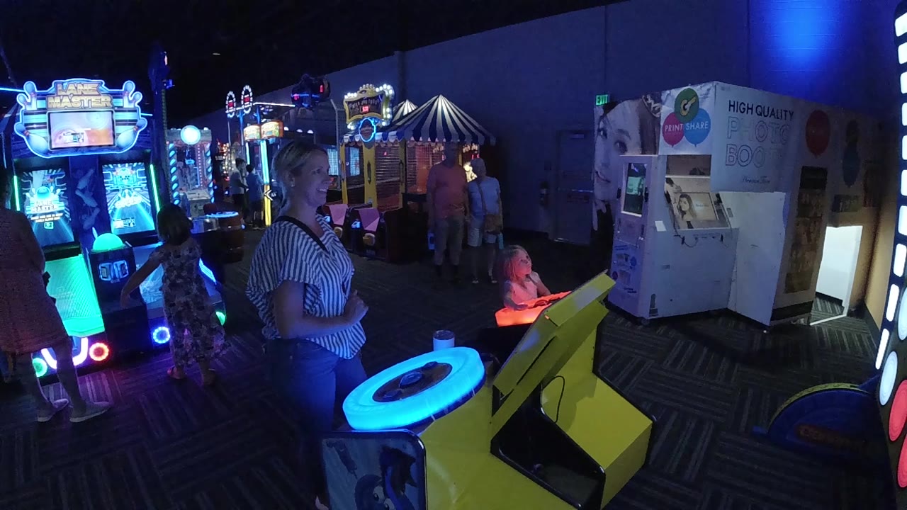 One second in, the woman remains near the glowing table as the child sits to the right and bright arcade cabinets shine in the background.