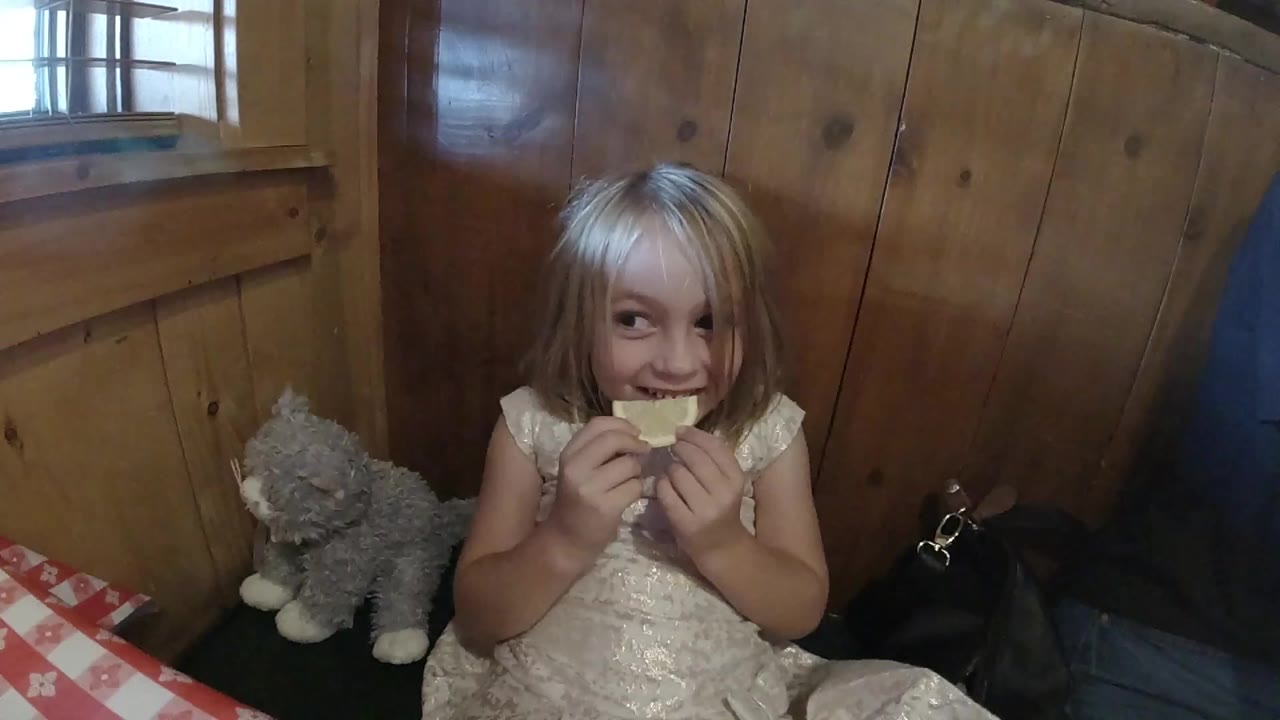 At the start of the Colorado clip, a blonde young girl sits in a wood-paneled interior holding an orange slice near her mouth with a gray stuffed animal beside her.