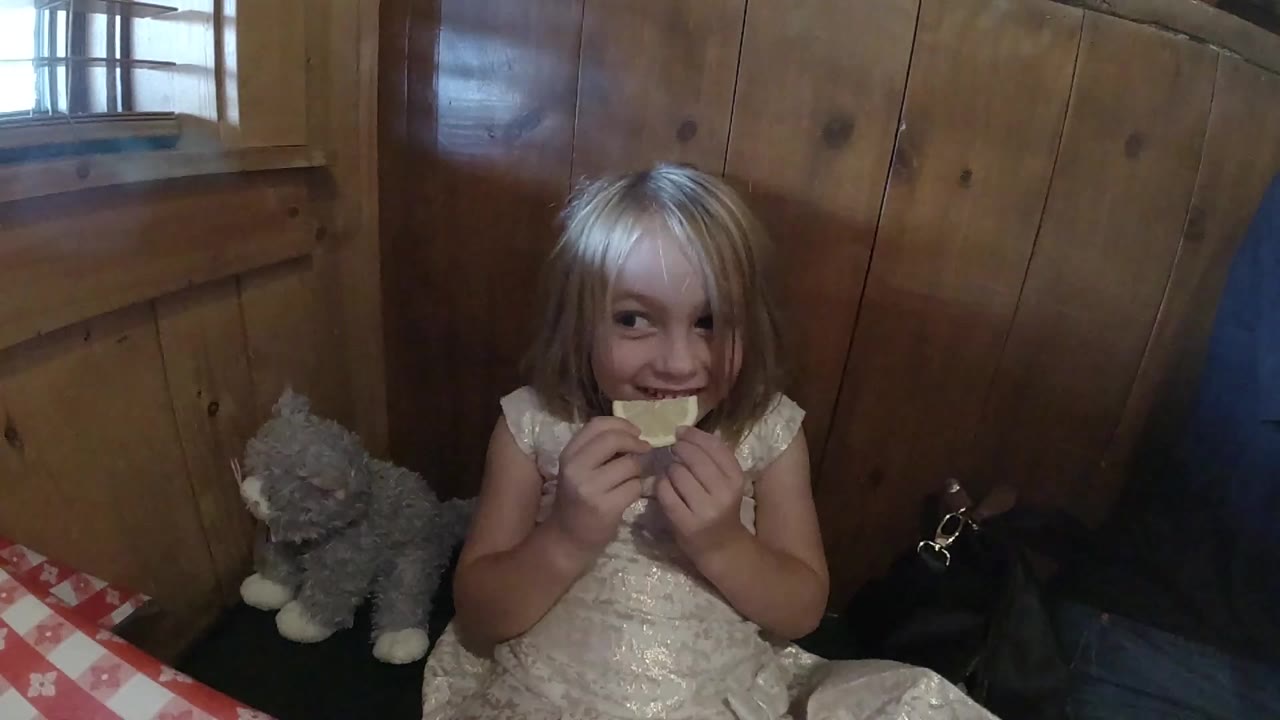 The girl faces forward in the Colorado indoor scene, gripping the orange slice with both hands while the stuffed animal remains on the seat to her left.