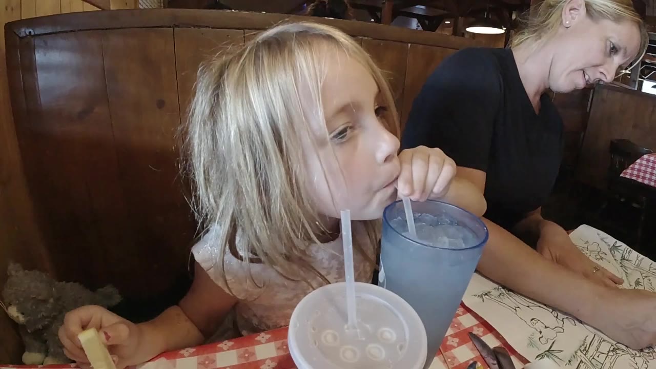 The girl keeps the straw at her mouth as the blue cup sits close to her face and the white lidded cup remains in the foreground.
