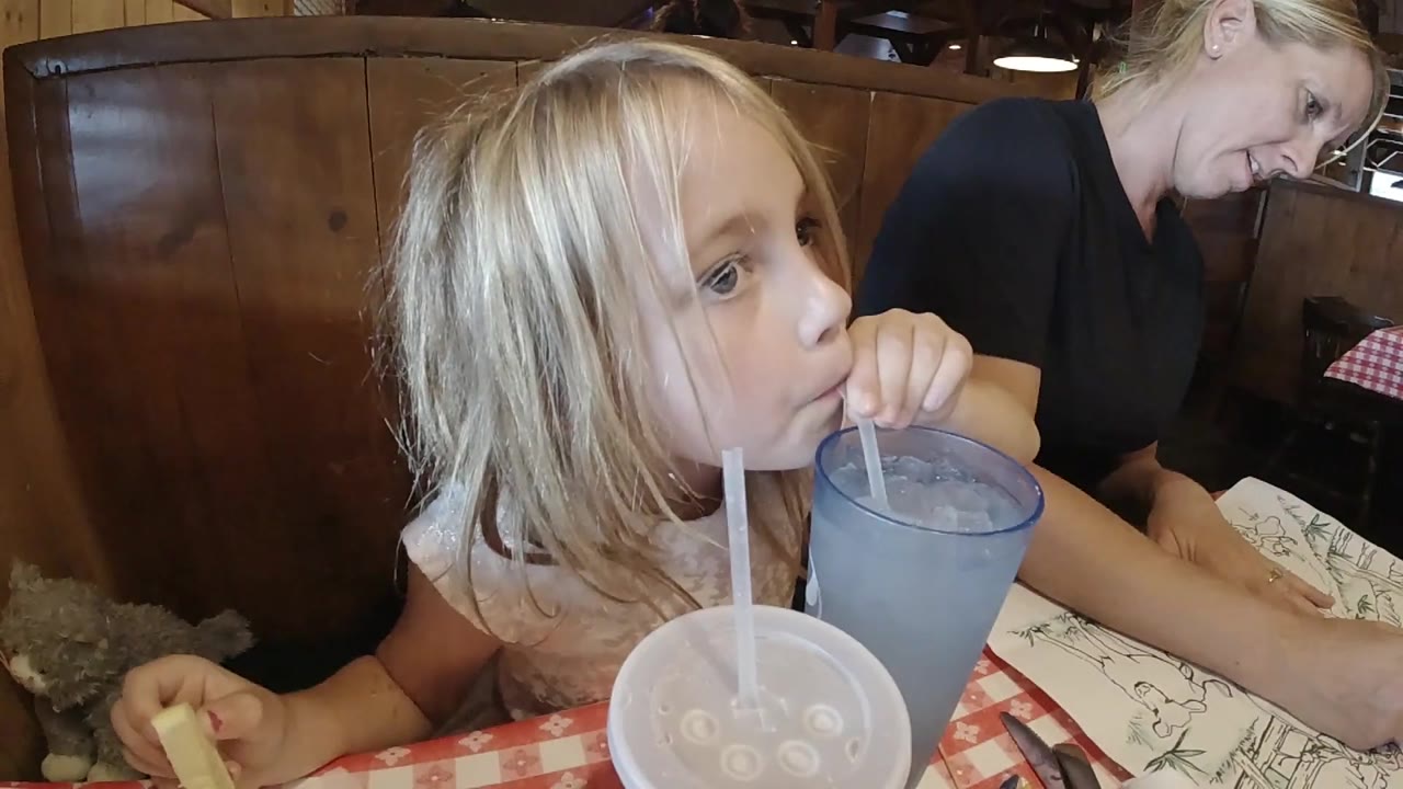 The child looks off to the right while continuing to sip from the blue cup, with the adult woman leaning over the table in the background.
