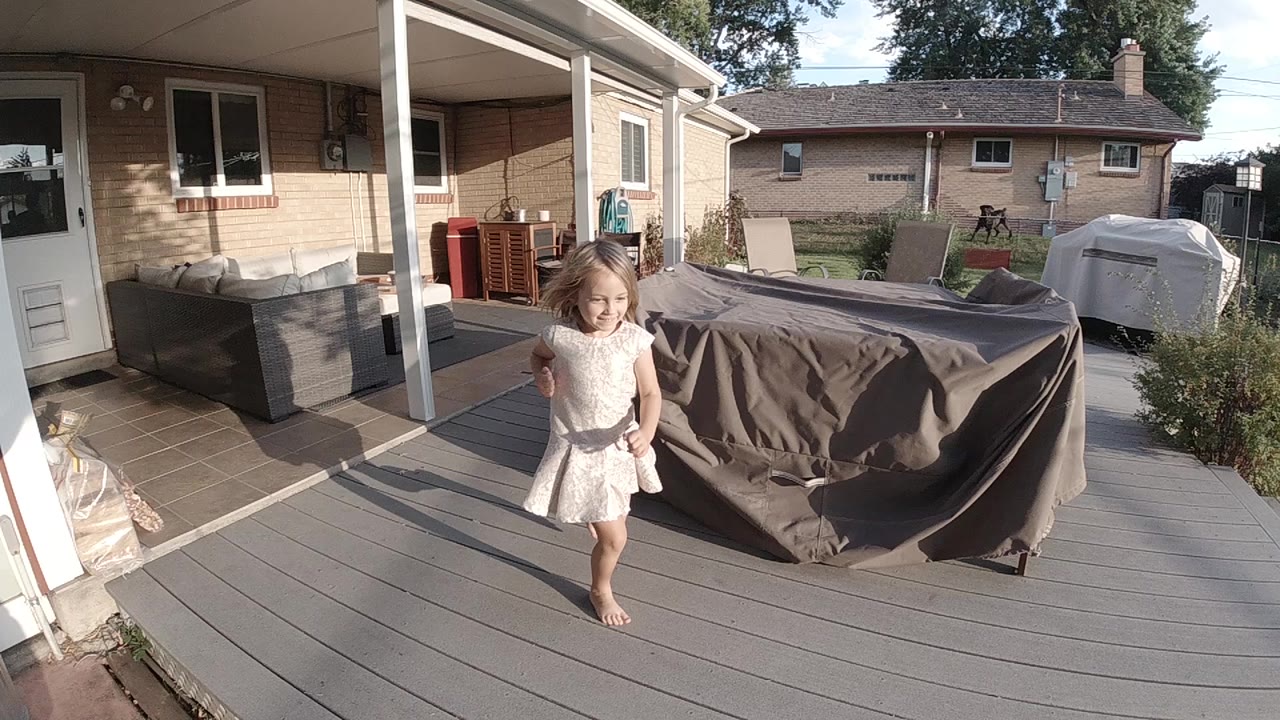 A young blonde girl in a light dress stands barefoot on a gray deck in Colorado, with a covered patio, outdoor furniture, and neighboring homes behind her.