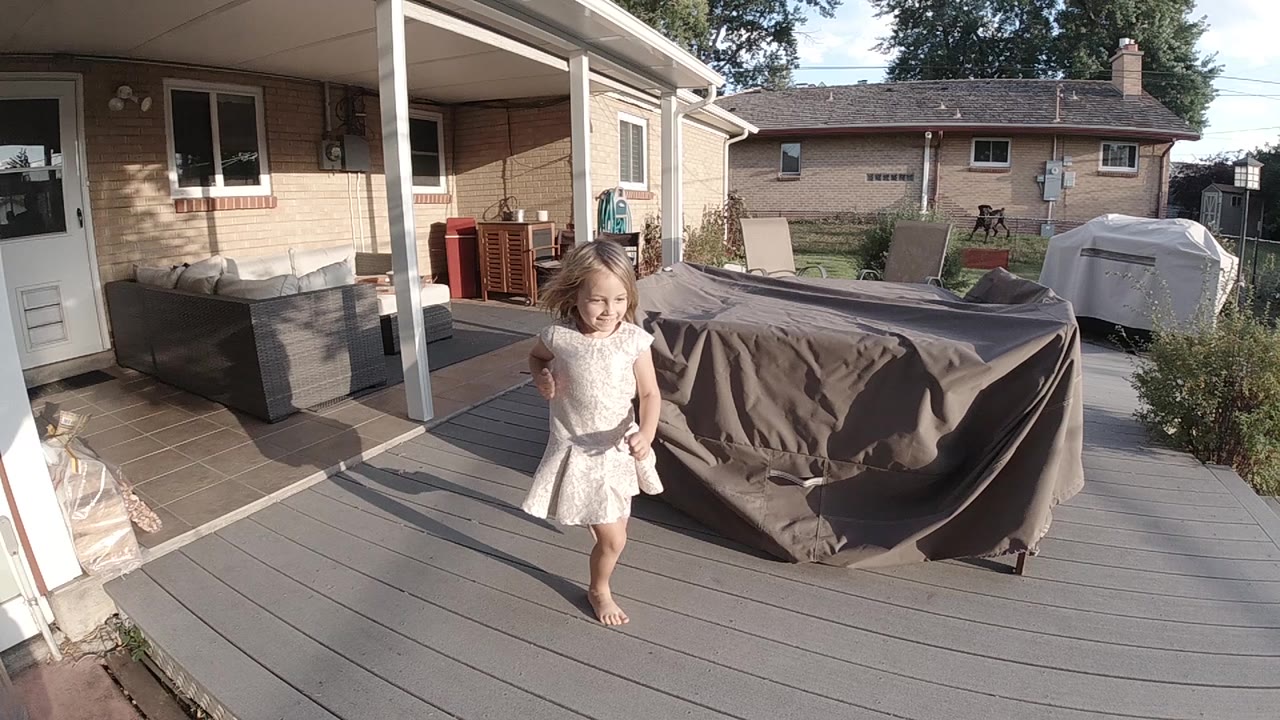 The girl starts forward across the backyard deck in Colorado, with her dress moving and the covered patio and draped outdoor furniture in the background.