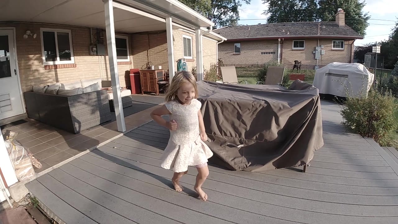 The child runs barefoot toward the camera across the deck, with sunlight hitting her dress and the backyard lawn and neighboring house visible beyond.