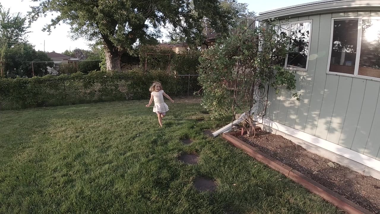The girl has moved closer through the Colorado yard, centered left of the pale building and framed by grass, stepping stones, shrubs, and a large tree.