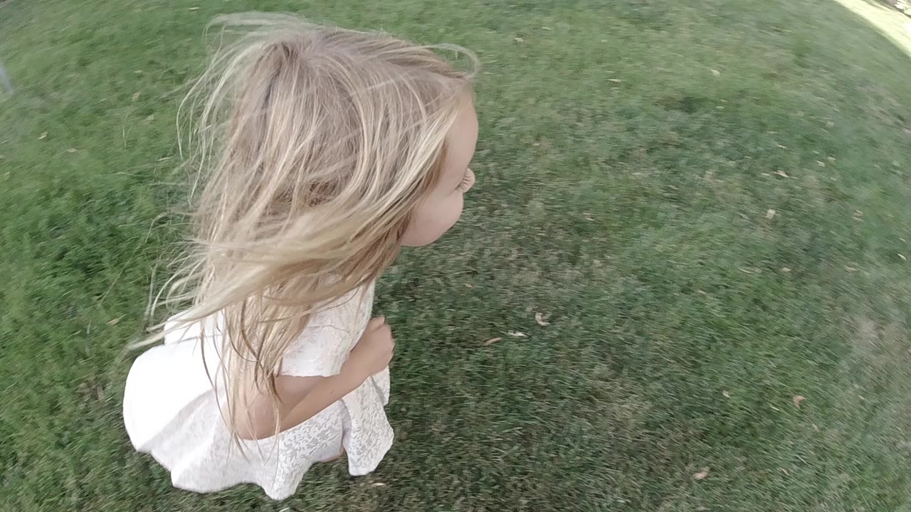 A blonde little girl in a white dress runs barefoot across green grass in Colorado, United States, seen from a high close angle at the start of the clip.