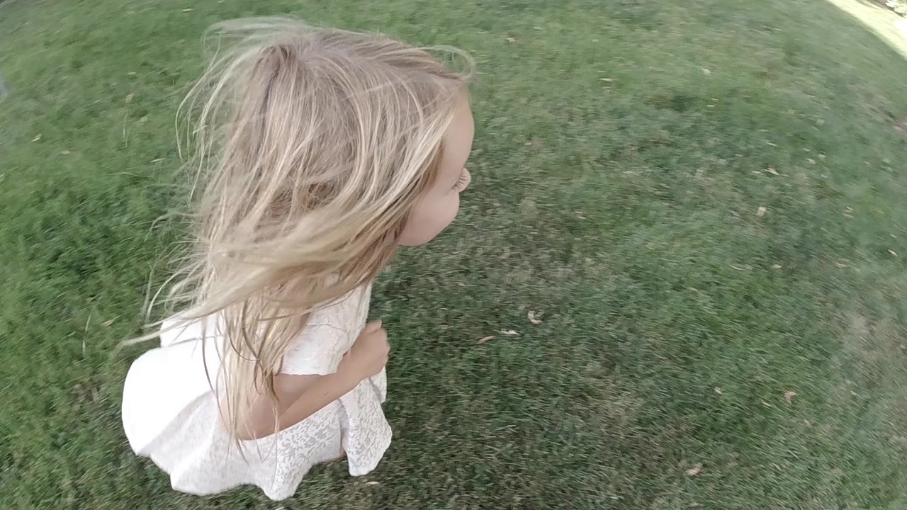 The little girl continues running over the grass, with her hair blown back and most of the frame filled by the lawn.