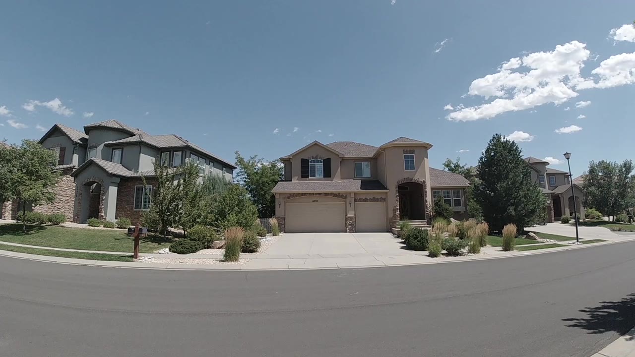 Early frame of a Colorado suburban street facing two-story homes, garages, trimmed lawns, shrubs, and a blue sky with no visible people.