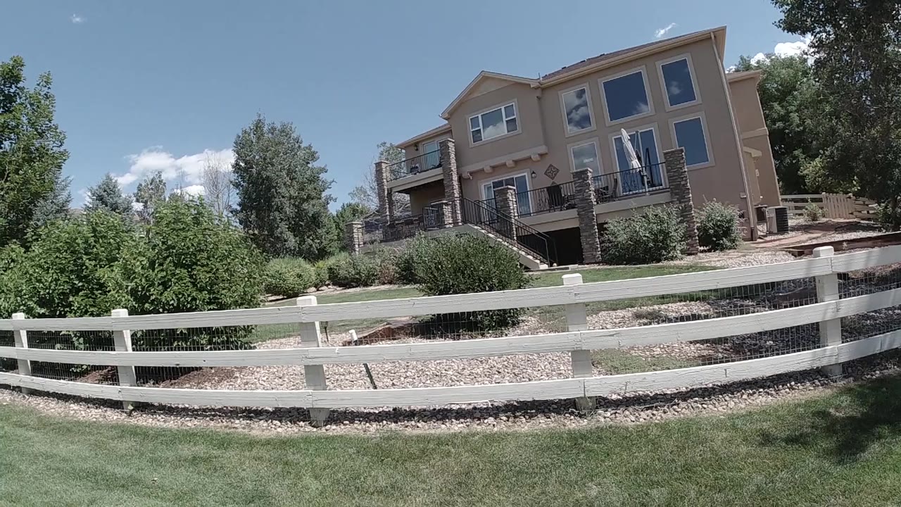 A slightly shifted wide view shows the white fence crossing the frame below the tan Colorado home, with shrubs and rock beds in bright sun and no visible people.
