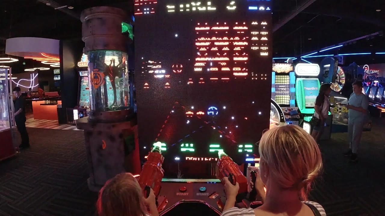 At 0:00, an adult and child in Colorado are seen from behind at a tall arcade game screen filled with red and green pixel enemies.