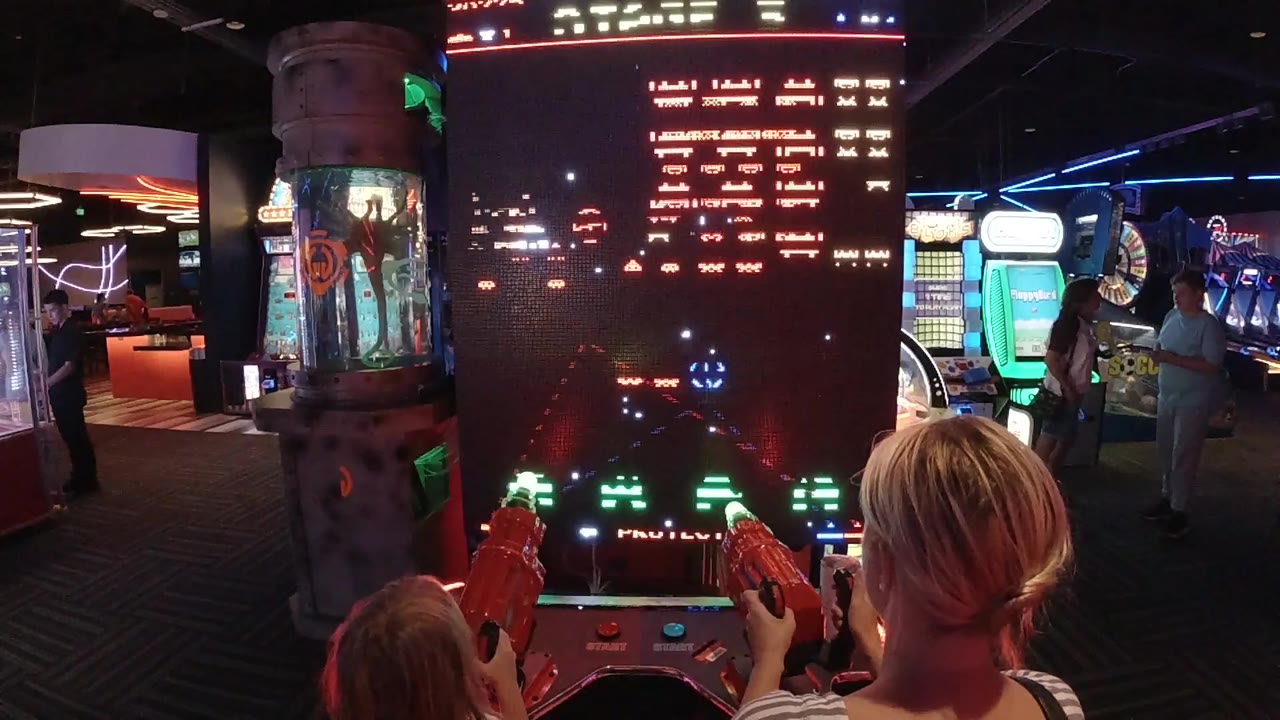 Two players face a tall arcade screen with bright game graphics.