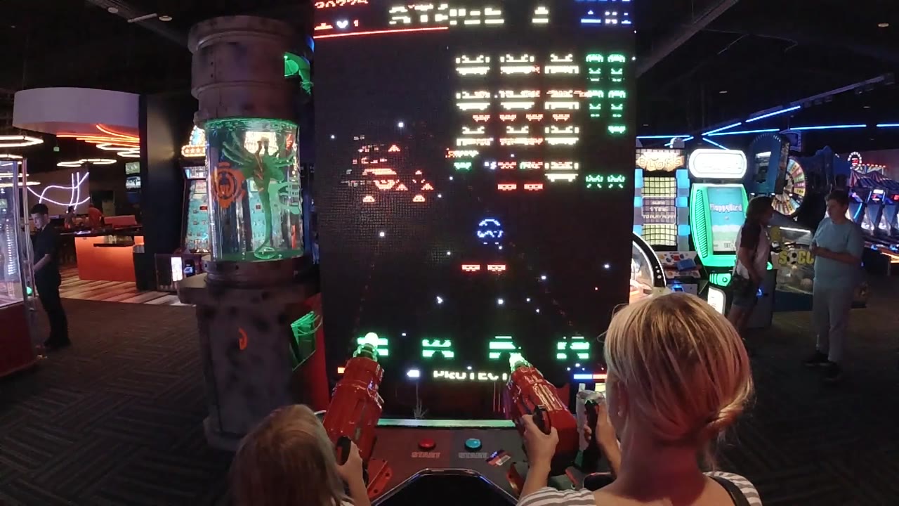 At 0:02, both players hold the red controls as the arcade game displays green, blue, and red rows against a black screen.