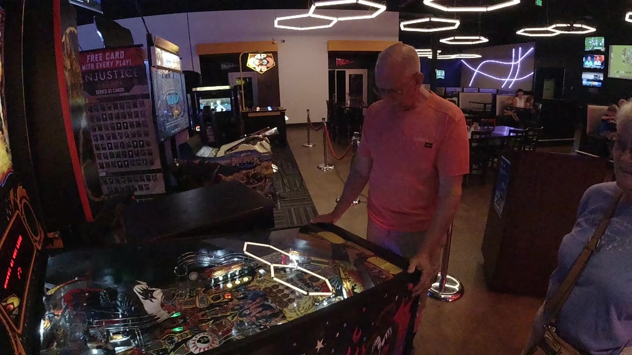 A bald man in a pink shirt stands over a lit pinball machine in a Colorado arcade room while another person is partly visible at the right edge.
