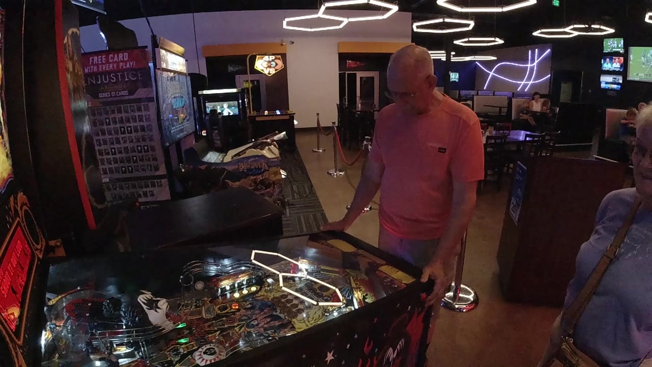 The player looks down at the pinball machine controls as ceiling ring lights reflect across the glass in the Colorado arcade interior.