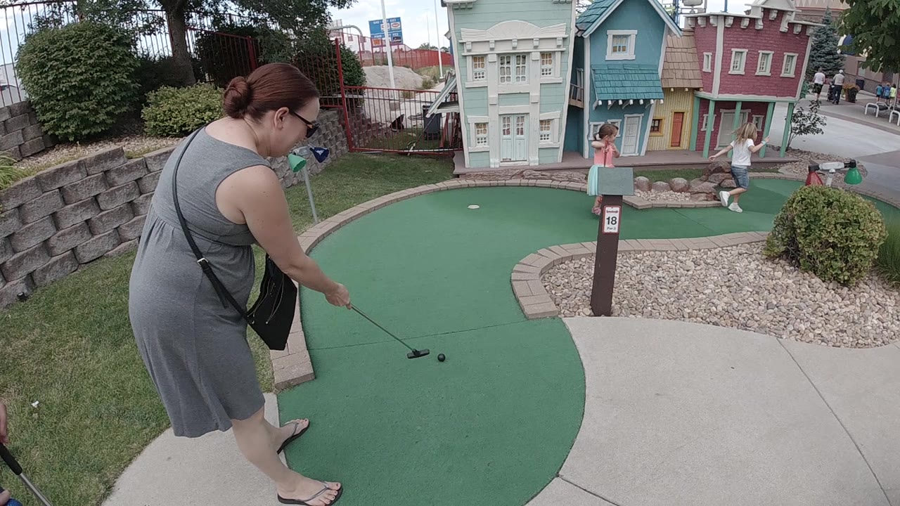 The woman stands over the ball with her putter angled forward while children and colorful facade buildings remain visible in the Denver course background.