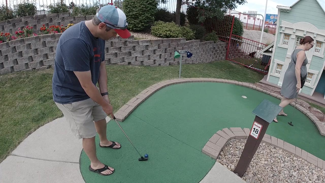 In Denver, a man in a cap stands over a putter on green mini golf turf while a woman appears near the painted building facade at the right edge.