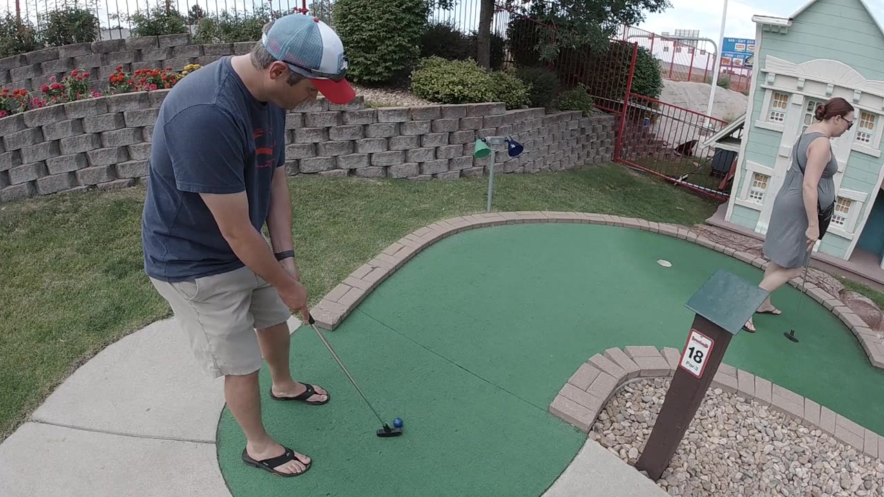 The man keeps his putter behind the ball on the green while the woman remains beside the miniature street-style building fronts in the background.