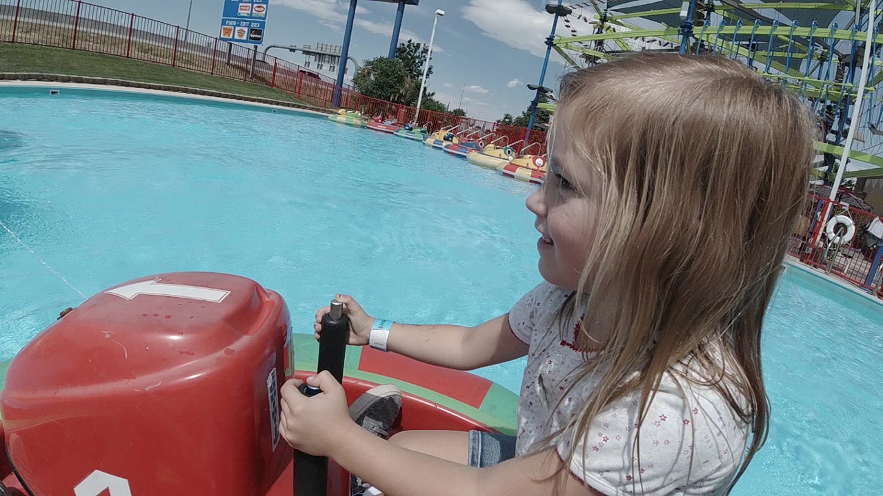 A young blonde girl in Denver sits close to the camera, holding black controls on a red bumper boat with blue pool water and fencing behind her.