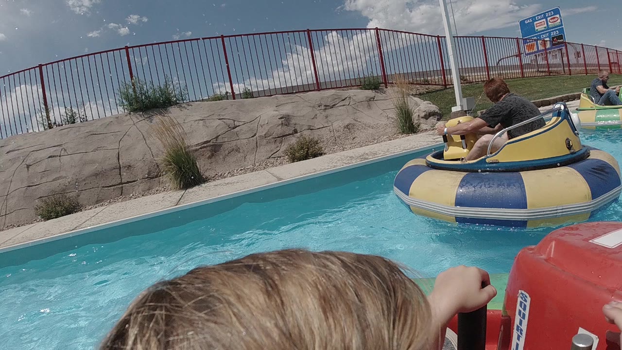 In Denver, a child's head and hand appear in the foreground of a red bumper boat while a yellow and blue boat floats ahead on bright blue pool water.