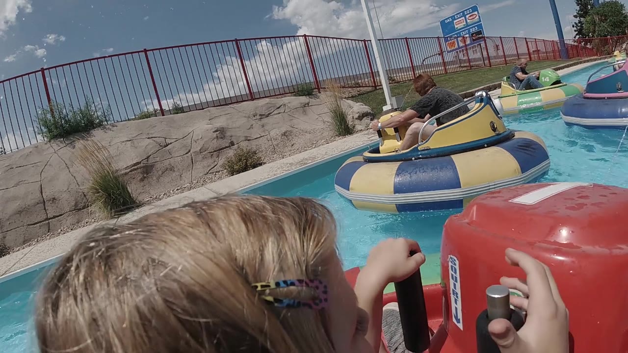 In Denver, a child with a hair clip grips the red bumper boat controls as yellow, blue, and green boats sit across the sunlit pool.