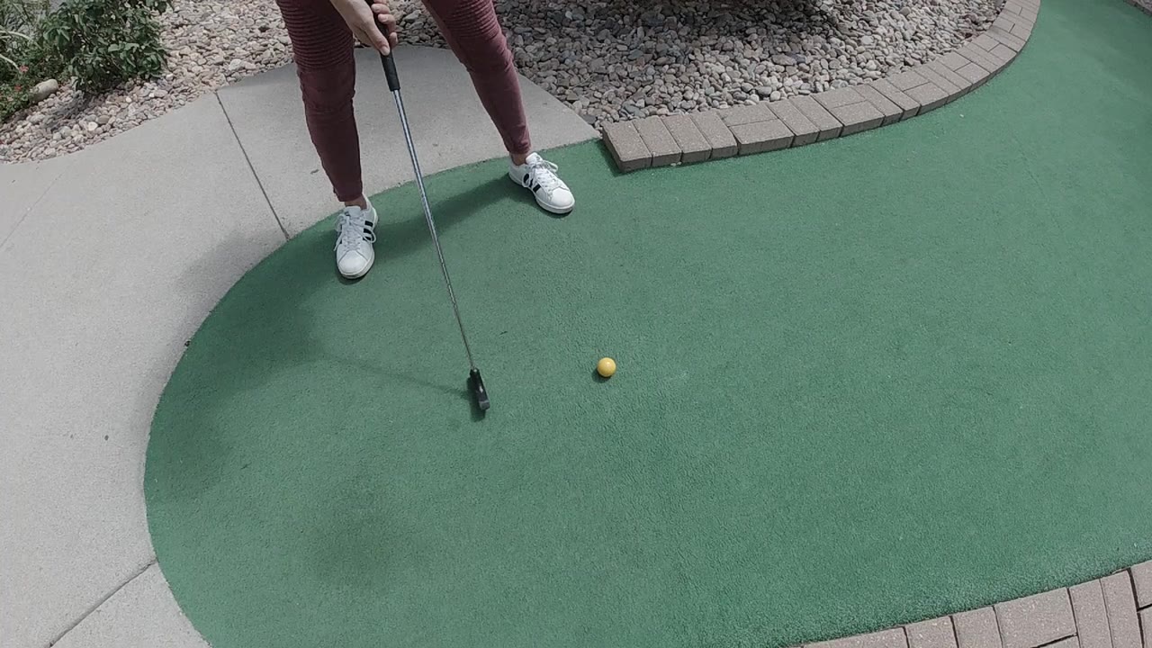 In Colorado, a golfer's lower legs and white shoes stand on a sunlit putting green as a black putter rests near a yellow ball.