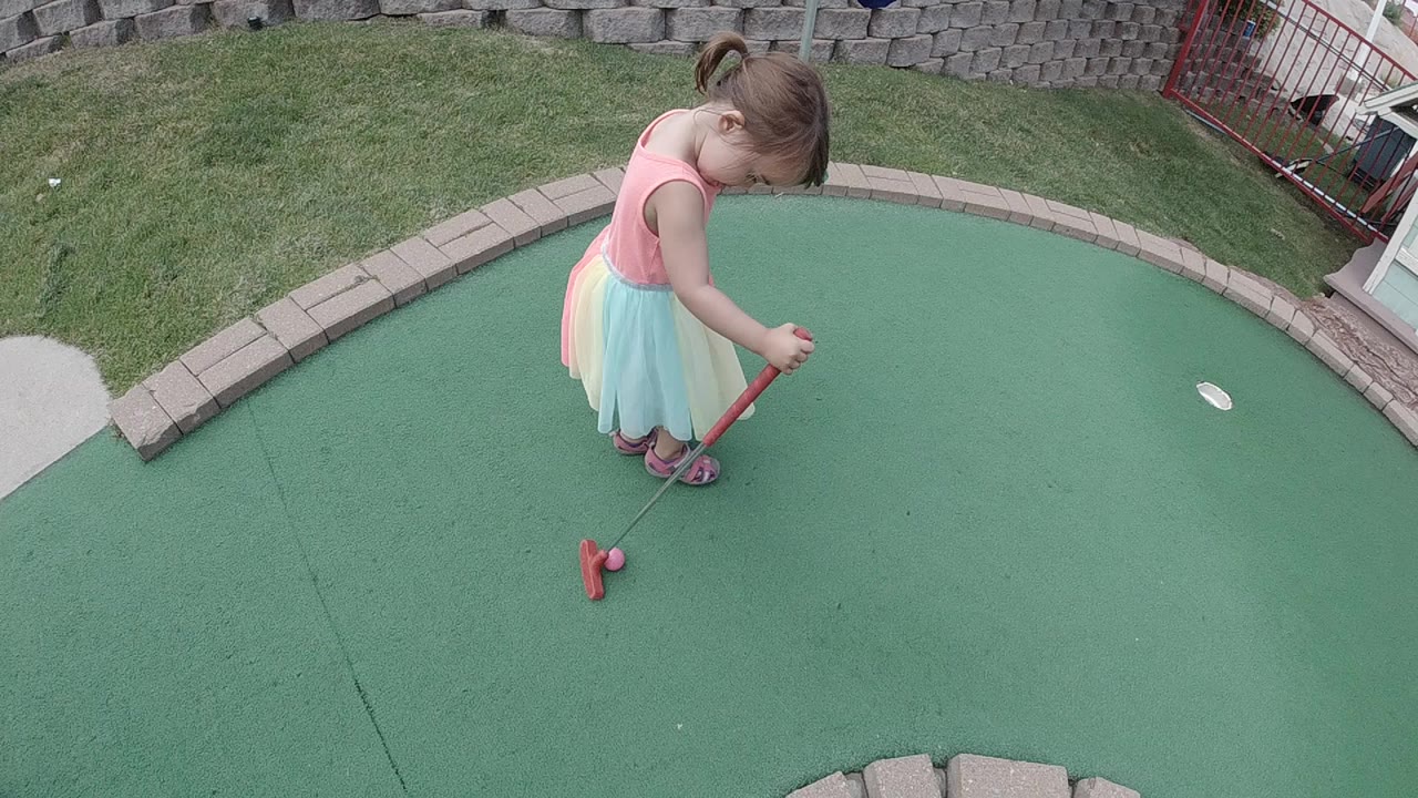 A young girl in a pastel dress looks down at a pink golf ball on a green mini golf surface in Colorado, with a red putter and curved stone border visible.