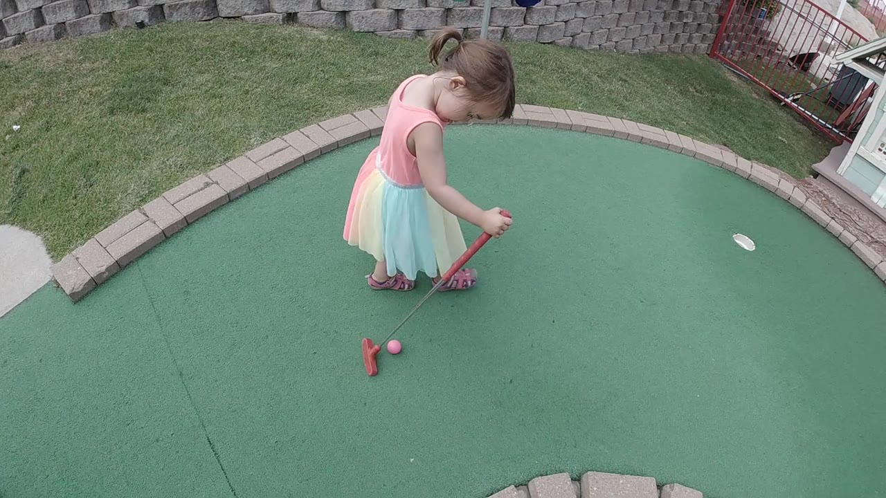 The child stands close to the ball with her putter lowered, while the hole sits off to the right on the green mini golf surface in Colorado.