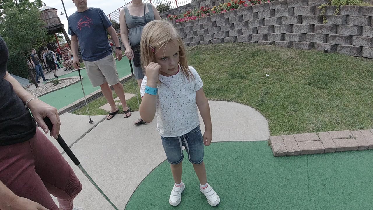 Young girl standing on green mini-golf turf near adults with putters.