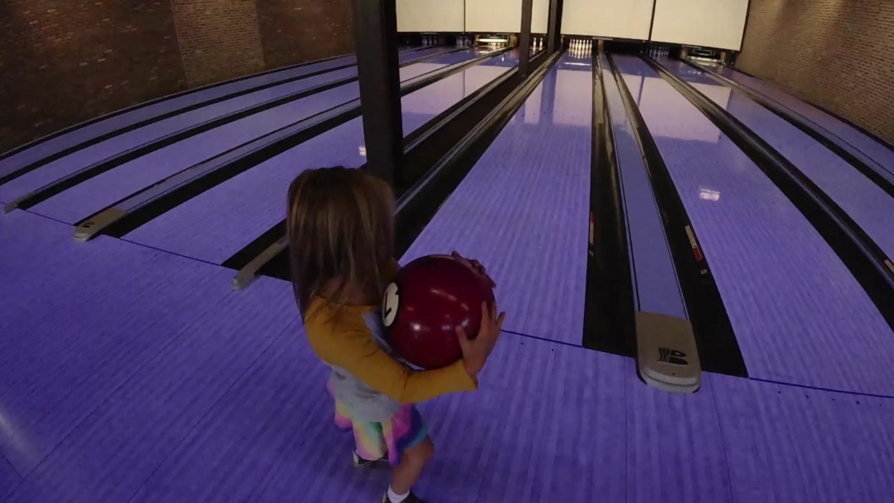 A young girl in a bright dress holds a red bowling ball near the start of a purple-lit bowling lane in Colorado.