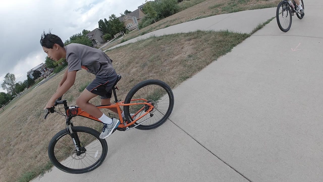 A boy on an orange bicycle rides close to the camera on a Denver path, with dry grass, cloudy sky, and distant homes behind him.