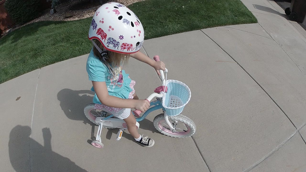 At the start of the Colorado clip, a young girl in a white patterned helmet rides a small white and pink bicycle on a sunlit concrete sidewalk beside green lawn.
