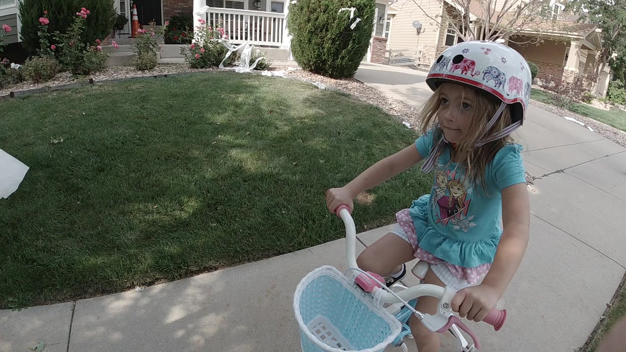 At the start of the Colorado clip, a young girl in a patterned helmet rides a small white bicycle beside a green lawn and residential sidewalk.