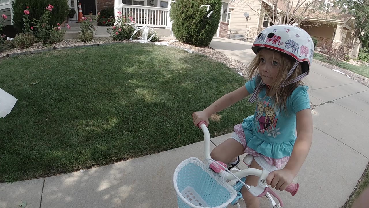 The girl continues along the sidewalk in Colorado, holding the handlebar while the lawn and porch railings fill the background.
