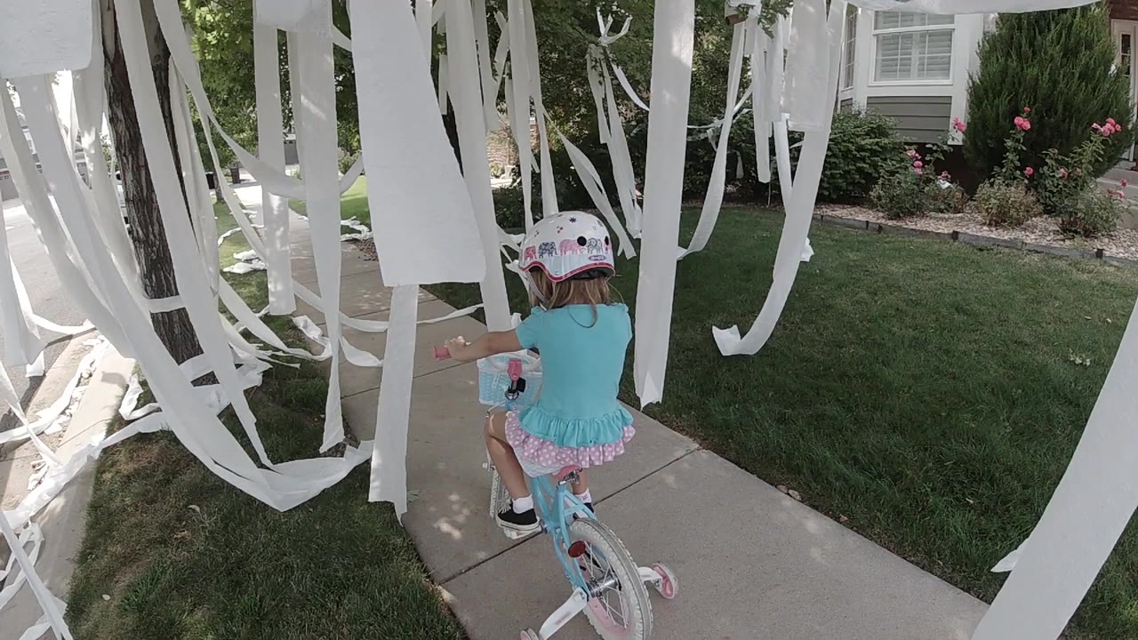 A helmeted girl in a turquoise top rides a pink bicycle along a sidewalk under hanging white strips beside a lawn and house windows.