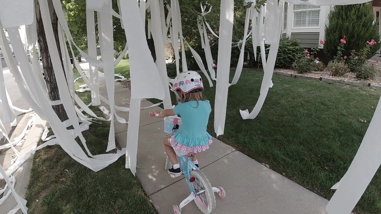A small child on a pink bicycle approaches a thicker cluster of white hanging ribbons with the house facade and flower bed beyond the sidewalk.