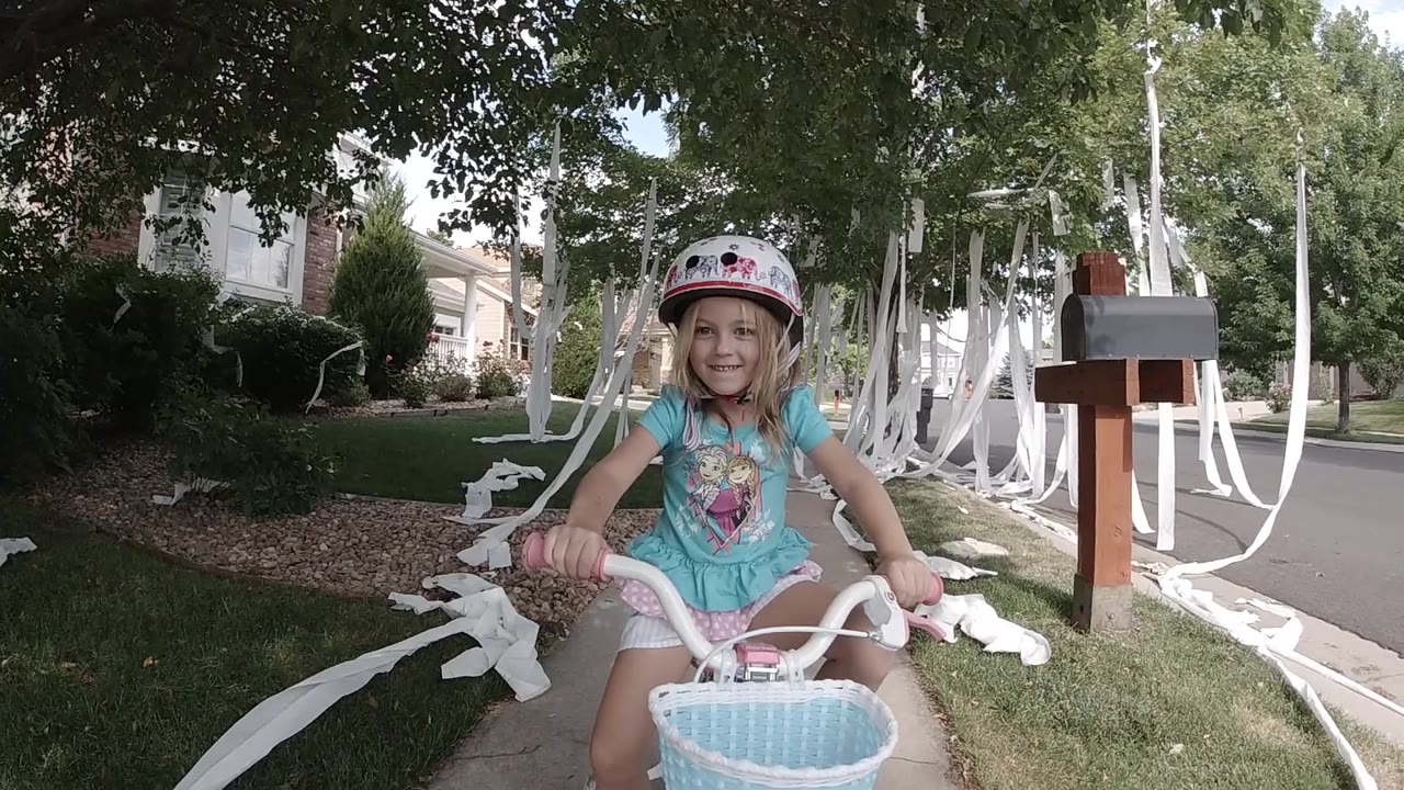 The girl continues smiling toward the camera while gripping the handlebars, with the decorated sidewalk and residential mailboxes still visible in Colorado.