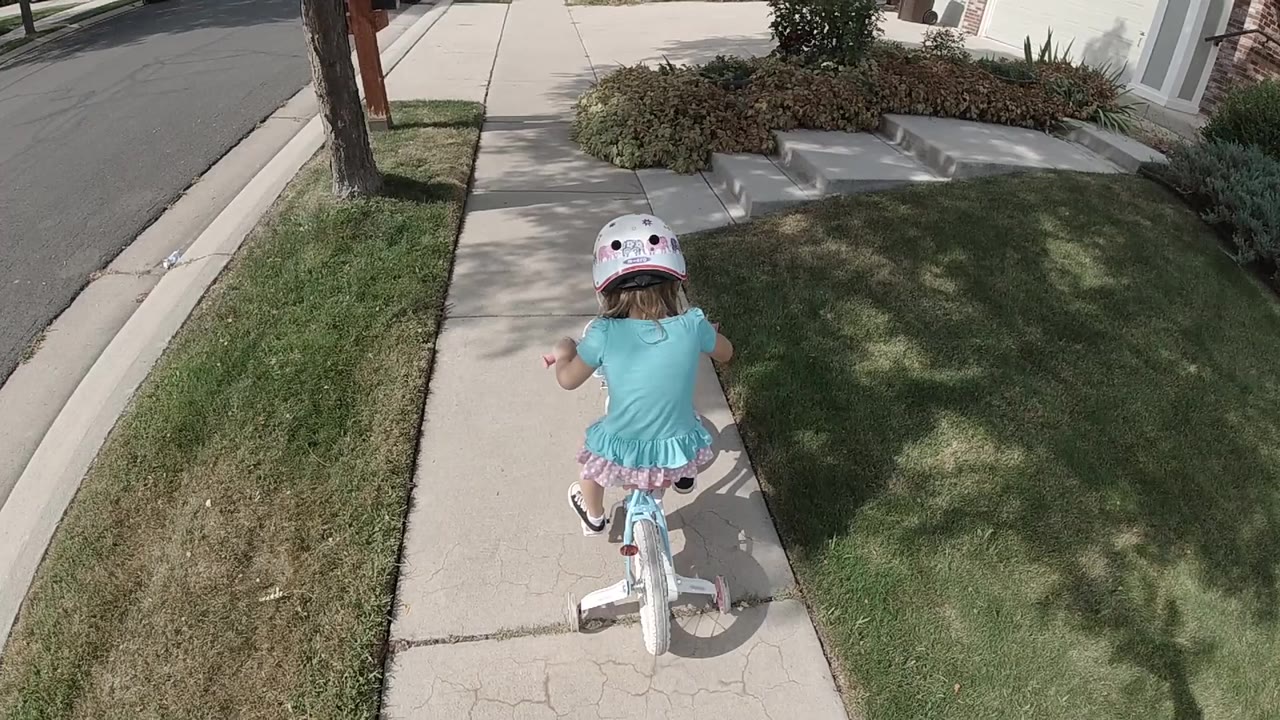 The child continues pedaling away from the camera, with a grass strip on the left, shaded lawn on the right, and residential sidewalk ahead in Colorado, United States.