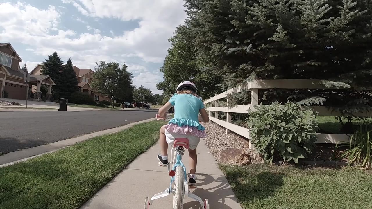 Little girl riding a bicycle away on a sidewalk beside a white fence.