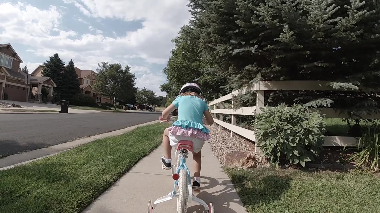 The rider remains centered on the sidewalk, pedaling away between the residential street on the left and the white fence line on the right in Colorado.
