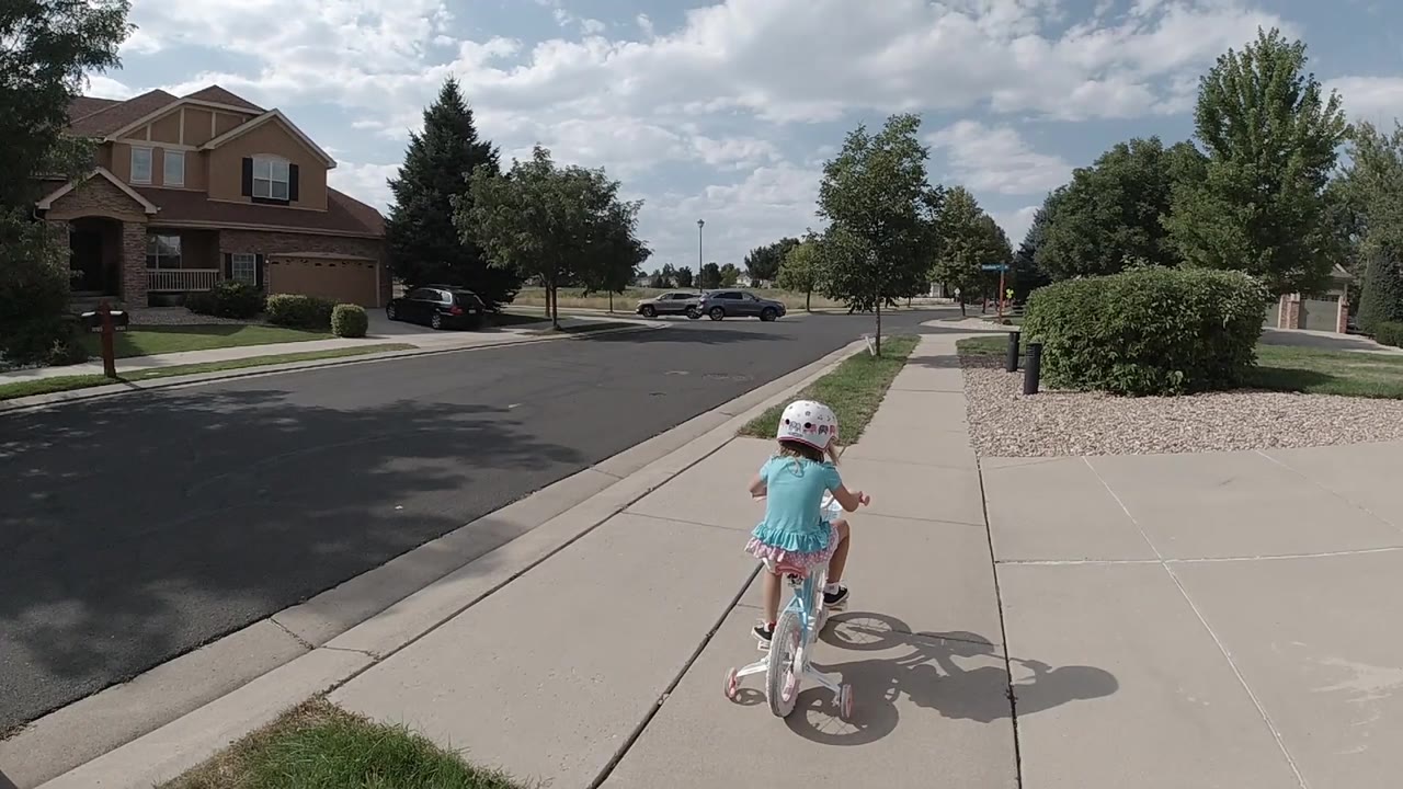 At 0:01, the helmeted girl pedals farther along the concrete path while parked cars and trimmed shrubs remain ahead in the Colorado neighborhood.